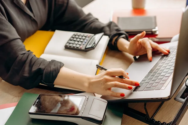 Close-up of a person using a laptop with a payment terminal and calculator on the desk. Zakelijke betaalrekening kiezen: eisen, functies en kosten